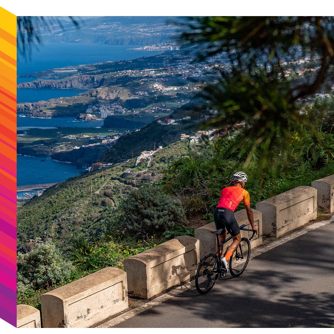 Rennradfahrer tragen das Luxa Magnetico Trikot aus der Fr&uuml;hjahr/Sommer-Kollektion 2023, w&auml;hrend im Hintergrund der Ozean und die Stadt Garachico zu sehen sind.